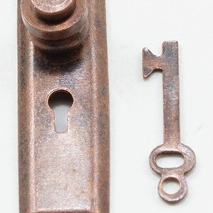 May include: A rectangular copper-toned metal escutcheon plate featuring a cylindrical knob and a central keyhole, accompanied by a matching small metal skeleton key. Both items exhibit a weathered, textured finish with dark brown patina accents.