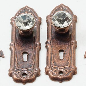 May include: Two copper-toned metal door plates with clear, faceted crystal knobs and keyhole cutouts, accompanied by two matching copper-toned skeleton keys. The rectangular plates feature decorative embossed edges and a weathered finish.