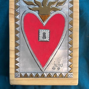 May include: A rectangular wooden plaque featuring a central red heart with a gold flame on top and a small keyhole icon in the middle. The heart is set against a silver background with a gold and silver triangular border. The word juju is signed.