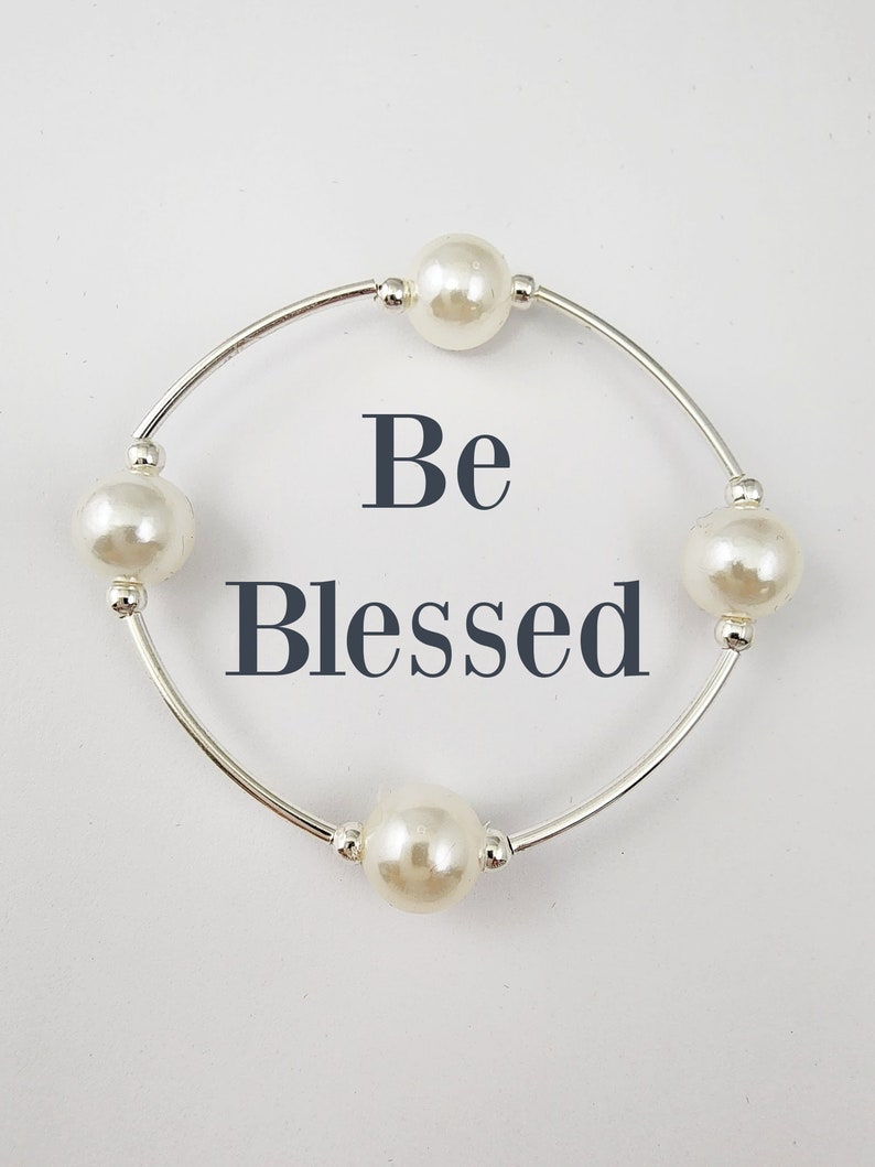May include: A silver bracelet with four white pearl beads and the words "Be Blessed" in black lettering.