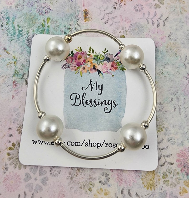 May include: A silver bangle bracelet with four white pearl beads. The bracelet is on a white card with the text "My Blessings" and a website address.