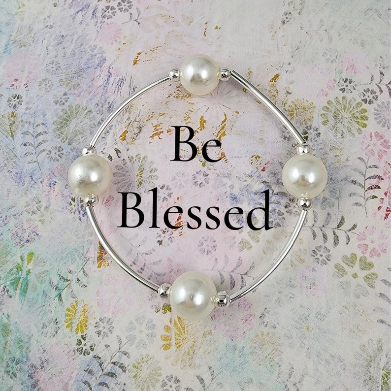 May include: A silver bracelet with four white pearl beads and the words "Be Blessed" on a white background.