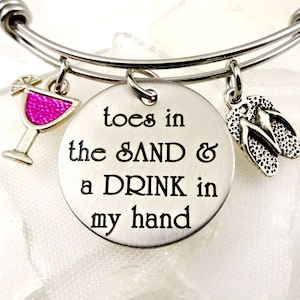 May include: Silver charm bracelet with a round charm that says "toes in the sand & a drink in my hand". The bracelet also has a charm of a pink cocktail glass and a charm of flip flops.