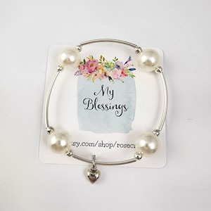 May include: A silver bangle bracelet with white faux pearl beads and a small silver heart charm. The bracelet has the text "My Blessings" on a card behind it.