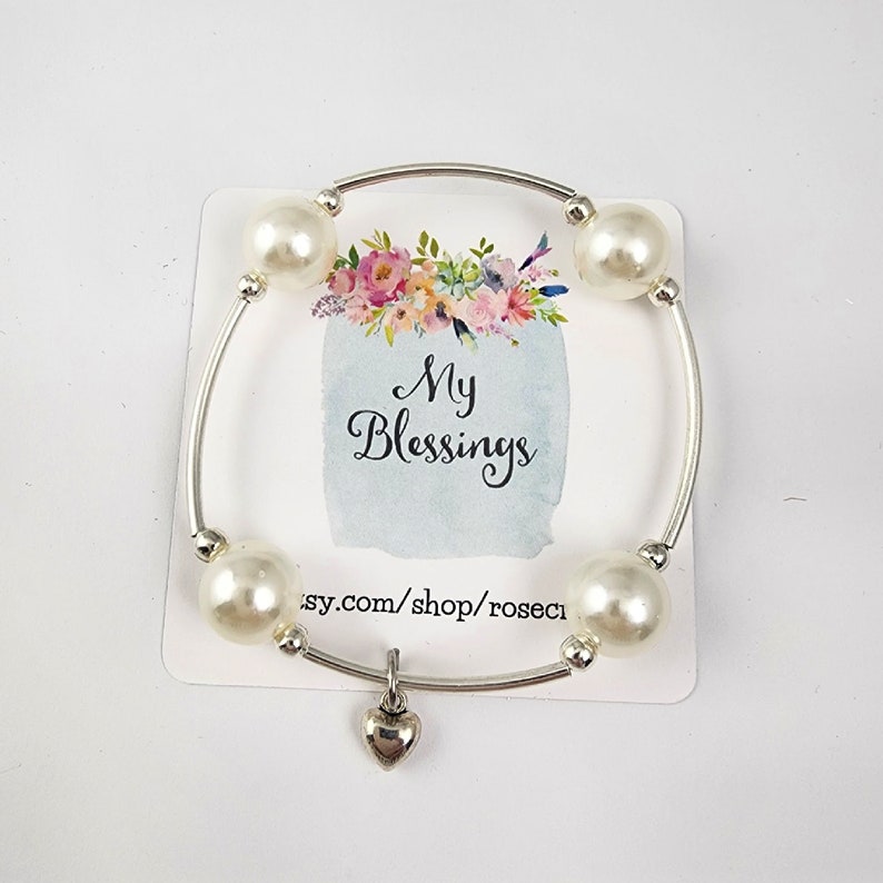 May include: A silver bangle bracelet with white faux pearl beads and a small silver heart charm. The bracelet has the text "My Blessings" on a card behind it.