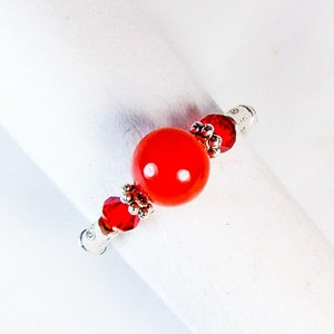 May include: A red beaded ring with a large, round, glossy red bead at the center. The ring features smaller red and silver beads on either side of the central bead. The ring is set against a white background.