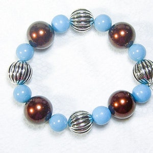 May include: A beaded bracelet featuring alternating light blue, brown, and silver beads. The brown beads have a glossy finish, while the silver beads are ribbed. The bracelet is strung on a clear elastic cord.