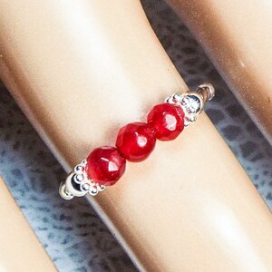 May include: A silver ring featuring three faceted, deep red beads. The beads are centered on the ring, with decorative silver accents on either side. The ring is shown on a light skin tone.