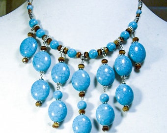 May include: A turquoise beaded necklace with a silver chain and brown accents. The necklace features large, oval turquoise beads that dangle from the main strand. The necklace is displayed on a white mannequin.