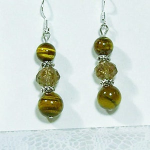 May include: A pair of dangle earrings featuring a combination of golden-brown tiger eye beads and faceted, light brown beads. The earrings have silver-colored metal hooks and decorative accents, creating a layered, elegant look.