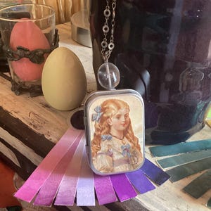 Puede incluir: Un collar de plata con una cuenta de cristal transparente y un pequeño medallón de metal con una ilustración vintage de una niña con cabello rubio que lleva un vestido blanco y cintas azules. El medallón está unido a una cadena de plata con una cuenta de cristal transparente. El medallón está rodeado de un arcoíris de tiras de tela púrpura, rosa y lavanda.