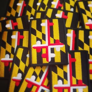 May include: A stack of black and yellow embroidered patches featuring the Maryland state flag. The flag is a black and gold checkered pattern with a red and white cross in the center.