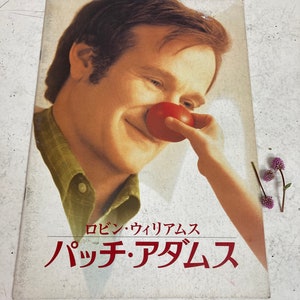 Könnte beinhalten: Ein Filmplakat mit Robin Williams, der einen roten Clownsnase trägt. Das Plakat ist für den Film "Patch Adams" und zeigt den Text "Robin Williams Patch Adams".