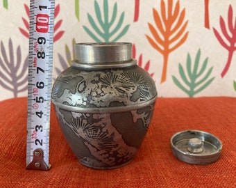 May include: A silver metal tea caddy with a lid and intricate floral designs. The caddy is sitting on a red surface.