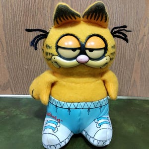 May include: A plush Garfield character toy featuring a yellow fuzzy body, black yarn whiskers, and a hard plastic face with half-closed eyes. The character wears light blue shorts with a printed Garfield logo and white sneakers with blue laces.