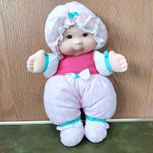 May include: A soft-bodied doll with a round face, brown eyes, and a small nose. The doll wears a pink and white floral bonnet with a teal bow, a bright pink ribbed top, and matching floral pants with teal ribbon accents at the wrists and ankles.