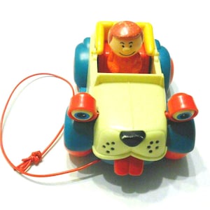 May include: A plastic pull-along toy shaped like a dog with a yellow body, blue wheels, and red accents. The toy features a small figure with a red head seated in the back, large googly eyes on the front, a black nose, and a red tongue.