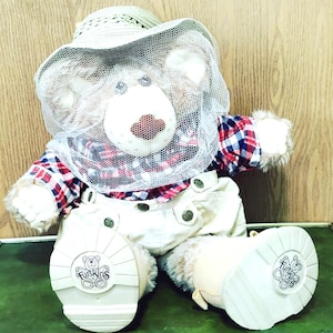 May include: A plush bear toy wearing a red, white, and blue plaid shirt, cream-colored overalls with metallic buttons, and a straw hat with a white mesh veil. The bear has light brown fur and white boots with the word Furskins printed on the soles.