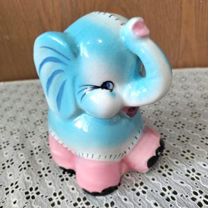 May include: A ceramic elephant figurine in a seated position. The elephant is primarily light blue with pink accents on the ears, skirt, and feet. The trunk is raised, and the eyes are detailed with black eyelashes.
