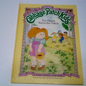 May include: A yellow and green book cover with the title "Cabbage Patch Kids" and the subtitle "The Shyest 'Kid in the 'Patch". The cover features a cartoon illustration of a Cabbage Patch Kid girl with red hair, wearing a purple dress and holding a basket of strawberries. The book is a Parker Brothers Story Book.