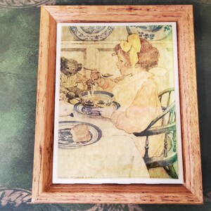 May include: A framed illustration of a girl and a bear sitting at a table, eating soup. The girl is wearing a yellow bow in her hair and a pink dress. The bear is wearing a brown vest. The illustration is set against a light background with a blue and white patterned tablecloth.