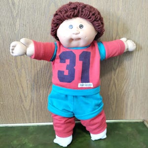 May include: A doll with a round face, dimples, and brown yarn hair. It wears a red long-sleeved shirt with teal accents and the number 31 in dark blue on the front, paired with matching teal shorts and red leggings. The doll has outstretched arms.
