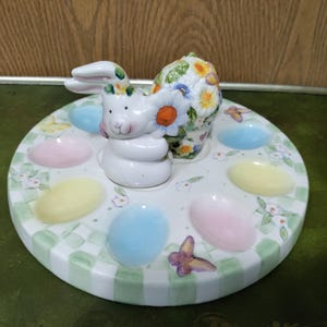 May include: A ceramic Easter egg holder featuring a white bunny with a floral basket. The round tray has pastel egg-shaped indentations in blue, pink, and yellow, with a green and white checkered border. Butterflies and flowers decorate the tray.