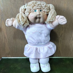 May include: A vintage Cabbage Patch Kids doll with blonde yarn hair styled in pigtails. The doll has blue eyes, rosy cheeks, and is wearing a pink dress with the Cabbage Patch Kids logo. White shoes complete the outfit.