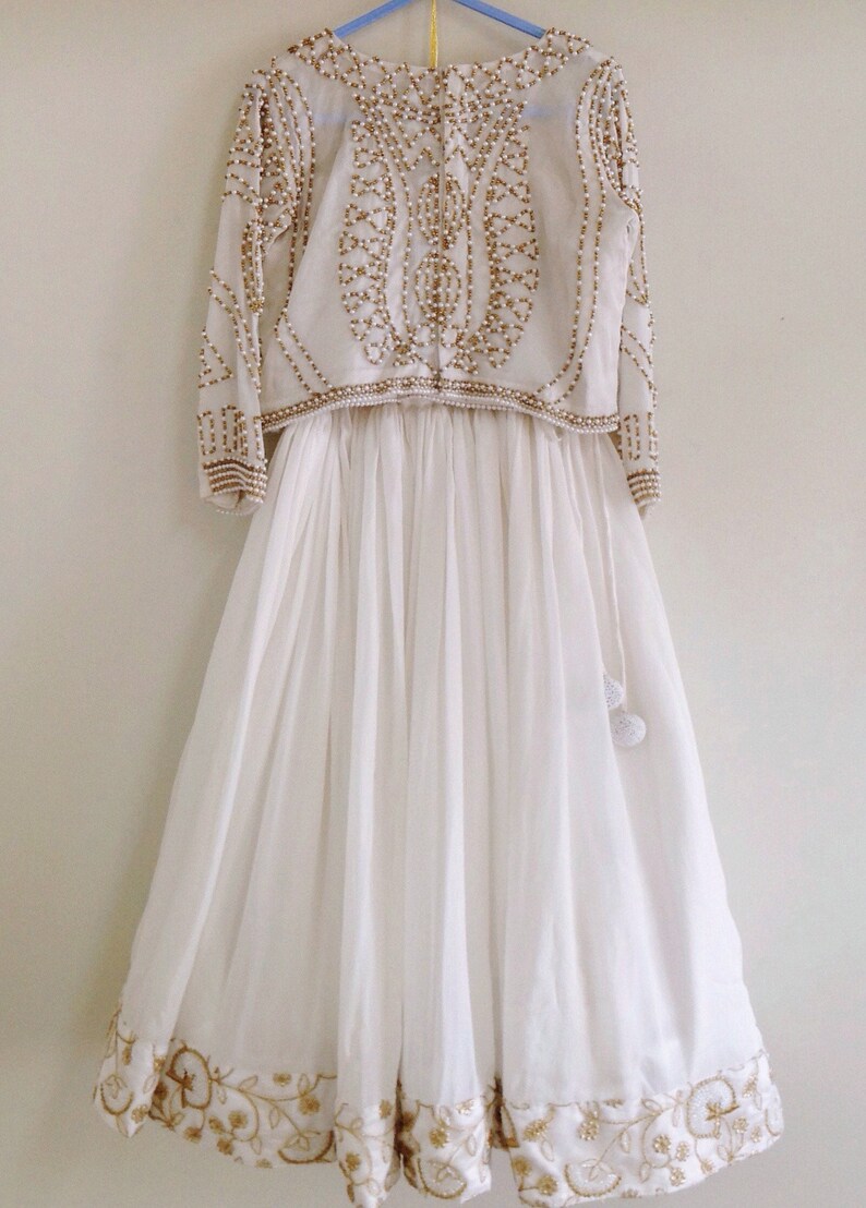May include: A white, floor-length skirt with gold embroidery and a matching white crop top with gold and pearl embellishments.