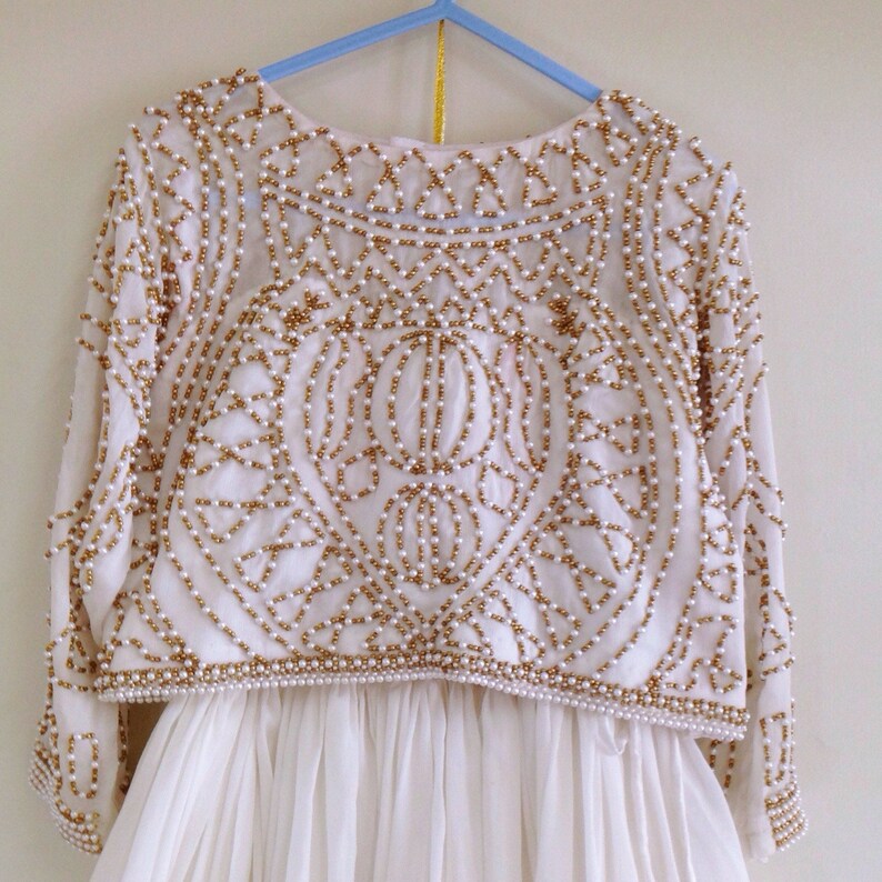 May include: A white dress with a long-sleeved, beaded top. The top features a geometric pattern with gold and white beads.