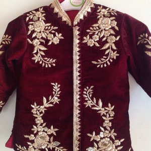 May include: Burgundy velvet jacket with gold embroidery of floral designs. The jacket has a mandarin collar and a button closure. The jacket is on a hanger with a tag that says "Lil Angels".