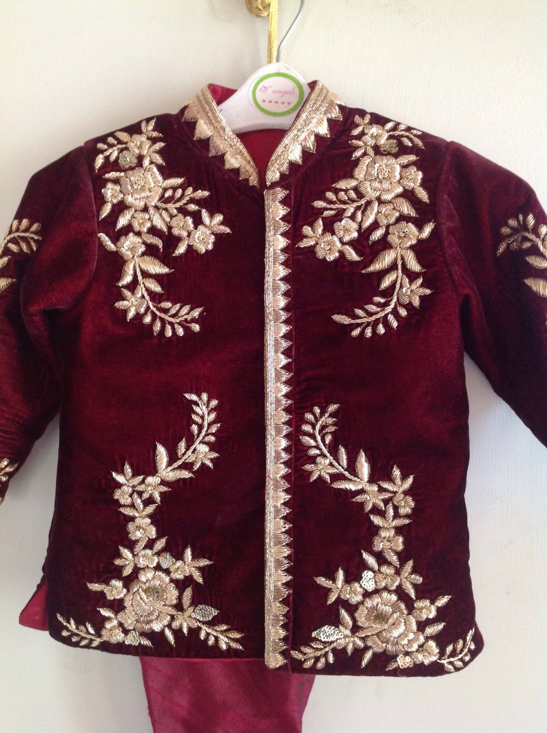 May include: Burgundy velvet jacket with gold embroidery of floral designs. The jacket has a mandarin collar and a button closure. The jacket is on a hanger with a tag that says "Lil Angels".