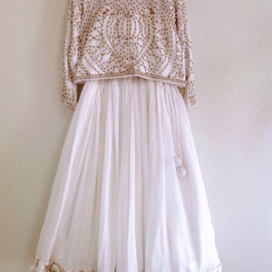 May include: A white, floor-length skirt with gold embroidery around the hem. The skirt is paired with a white crop top with gold and pearl beading.