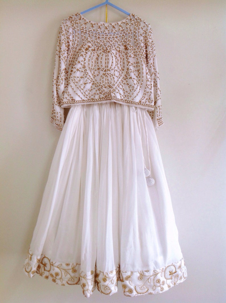May include: A white, floor-length skirt with gold embroidery around the hem. The skirt is paired with a white crop top with gold and pearl beading.
