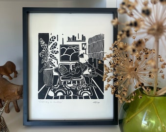 May include: Black and white linocut print of a person relaxing in a chair with a book. The print is framed in black and has the text "Especially on Sundays" at the bottom.