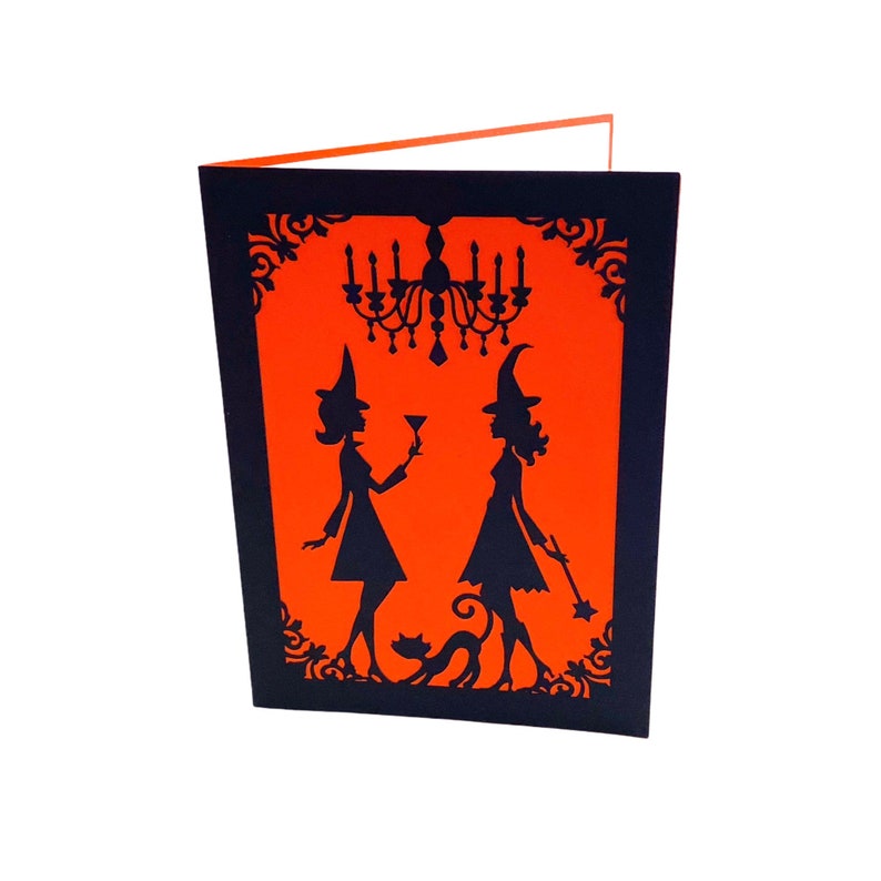 Pu&ograve; includere: Un biglietto di Halloween nero e arancione con un design pop-up. Due streghe sono in silhouette su uno sfondo arancione, alzando un bicchiere per un brindisi. Un gatto nero &egrave; seduto sotto di loro. Un lampadario pende sopra le streghe.