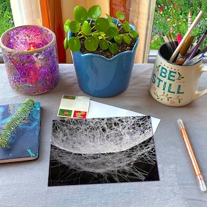 May include: A still life arrangement featuring a mosaic candle holder, a blue potted plant, and a mug with the words "BE STILL." Also present are a notebook, a pen, and a black and white photograph.