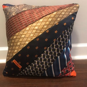 May include: A decorative pillow made from repurposed neckties. The pillow features a patchwork design with diagonal stripes of various patterns and colors, including paisley, diamonds, and stripes. The color palette includes shades of red, gold, navy, and orange.