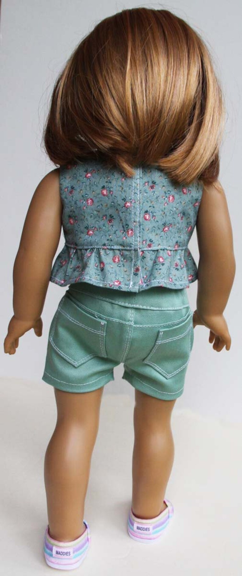 Peut inclure: Une poup&eacute;e portant un haut sans manches &agrave; imprim&eacute; floral vert avec un ourlet &agrave; volants et un short en jean vert. La poup&eacute;e porte des baskets ray&eacute;es arc-en-ciel avec le mot "Maddies" dessus.