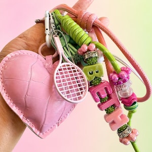 May include: A pink heart-shaped keychain with a textured surface, a white tennis racket charm, and a beaded charm spelling "Beth". The keychain also features green and pink accents.