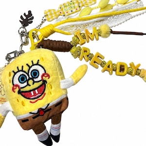 May include: A plush SpongeBob SquarePants keychain featuring a yellow character with blue eyes and a wide smile. The accessory includes yellow and white ribbons, a brown cord, and yellow letter beads spelling out I'M READY, plus a candy charm.