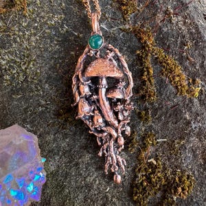 Puede incluir: Un collar con colgante en tono cobre que presenta un diseño detallado de setas y enredaderas con una pequeña piedra preciosa verde en la parte superior. El colgante cuelga de una cadena de cobre a juego, mostrando texturas intrincadas y elementos orgánicos.