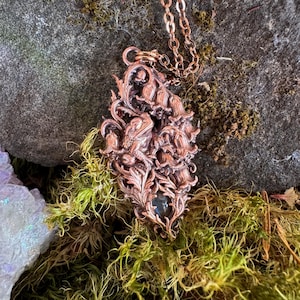 Puede incluir: Un collar con colgante de color cobre que presenta un diseño tridimensional detallado de una rana entre motivos florales y de hojas. El metal tiene un acabado oscuro y oxidado, y el colgante cuelga de una cadena de cobre a juego.