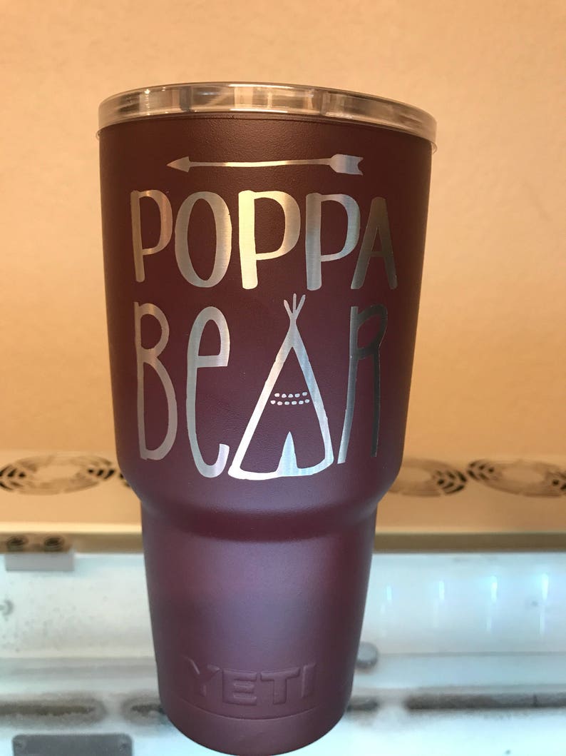 May include: A maroon Yeti tumbler with a silver design that says "Poppa Bear" with an arrow and a teepee graphic.