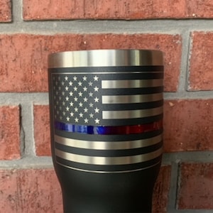 May include: Black stainless steel tumbler with a laser-etched American flag design. The flag has a thin blue line and a thin red line across the middle.