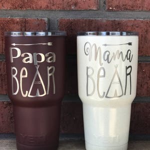 May include: Two stainless steel tumblers with lids. The tumbler on the left is brown with the text "Papa Bear" and a teepee design. The tumbler on the right is white with the text "Mama Bear" and a teepee design.
