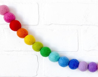 Puede incluir: Una guirnalda de bolas de fieltro de colores del arcoíris, incluyendo rojo, naranja, amarillo, verde, azul y morado. La guirnalda está colgada contra una pared de ladrillos blancos, creando una decoración vibrante y alegre.