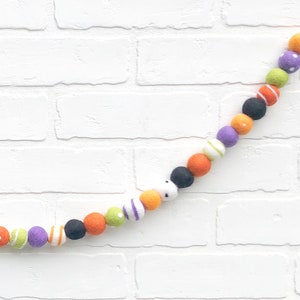 May include: A colorful Halloween garland featuring felt balls in orange, black, purple, green, and white. The balls are decorated with stripes, polka dots, and other patterns. The garland is strung on a white string and is displayed against a white brick wall.