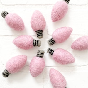 May include: Pink felt lightbulb ornaments with black and white stripes. The ornaments are arranged in a starburst pattern on a white brick background.