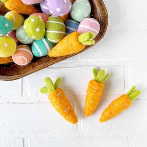May include: A wooden bowl filled with felt Easter eggs in various colors and patterns, including pink, purple, green, blue, and yellow. There are also three felt carrots with green tops.